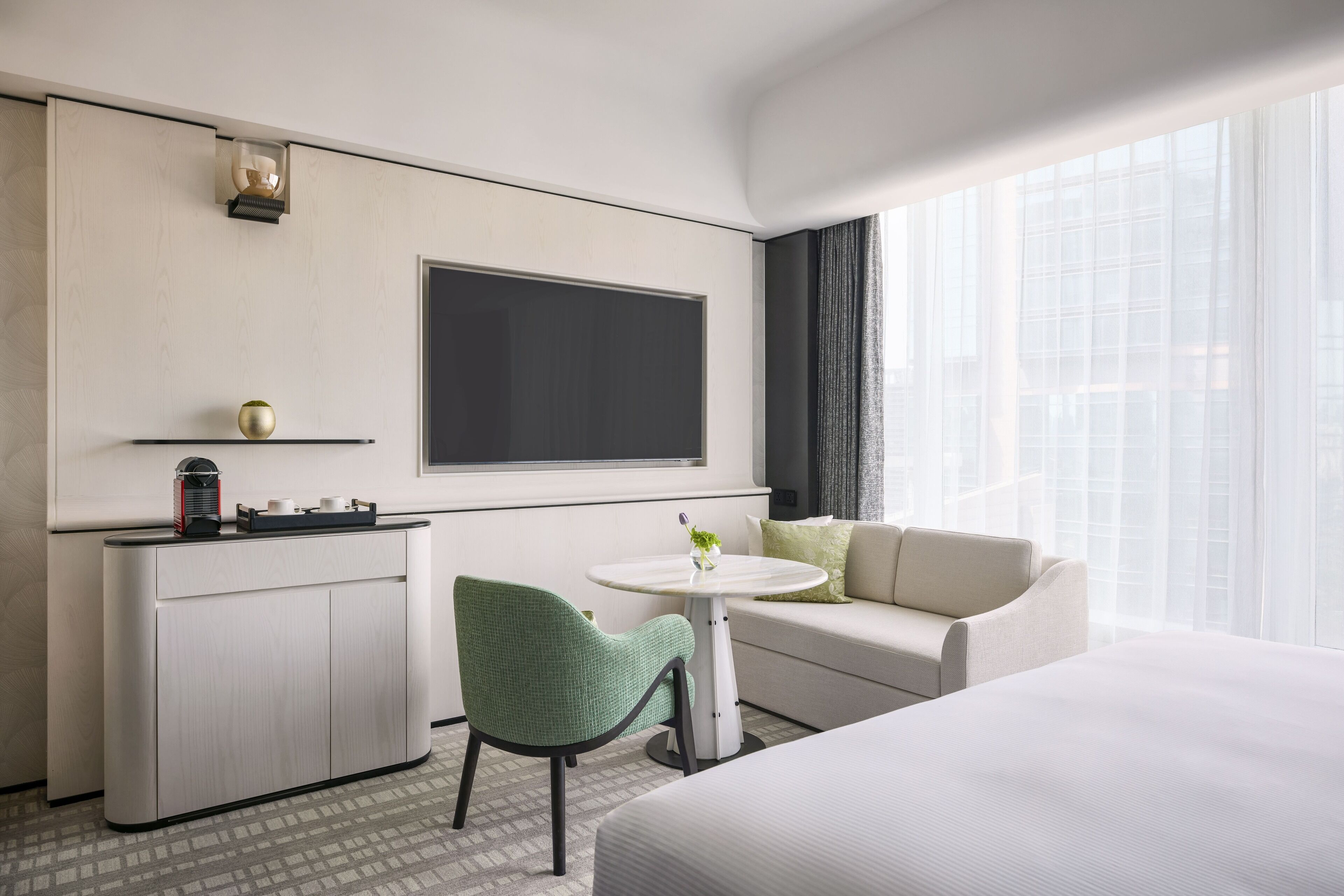 A hotel room with a bed, a sofa, a small table with a vase, a green chair, a minibar, and a large window with curtains.