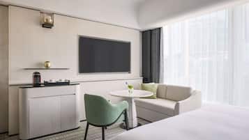A hotel room with a bed, a sofa, a small table with a vase, a green chair, a minibar, and a large window with curtains.