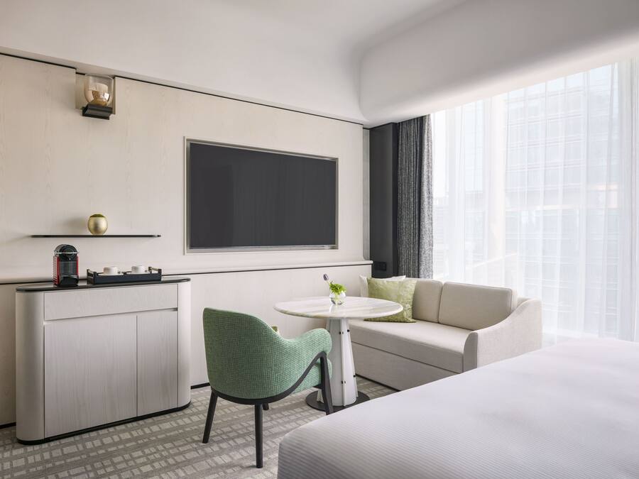 A hotel room with a bed, a sofa, a small table with a vase, a green chair, a minibar, and a large window with curtains.