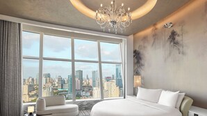 Ein modernes Hotelzimmer mit einem großen Bett, einem runden Sessel und Blick auf eine Stadtlandschaft durch große Fenster.