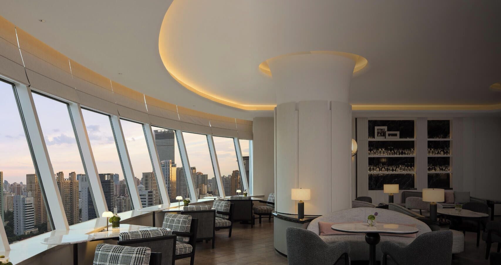A high-rise hotel room with a panoramic city view, a round dining table, and a bar area with shelves of glasses.
