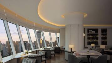 A high-rise hotel room with a panoramic city view, a round dining table, and a bar area with shelves of glasses.