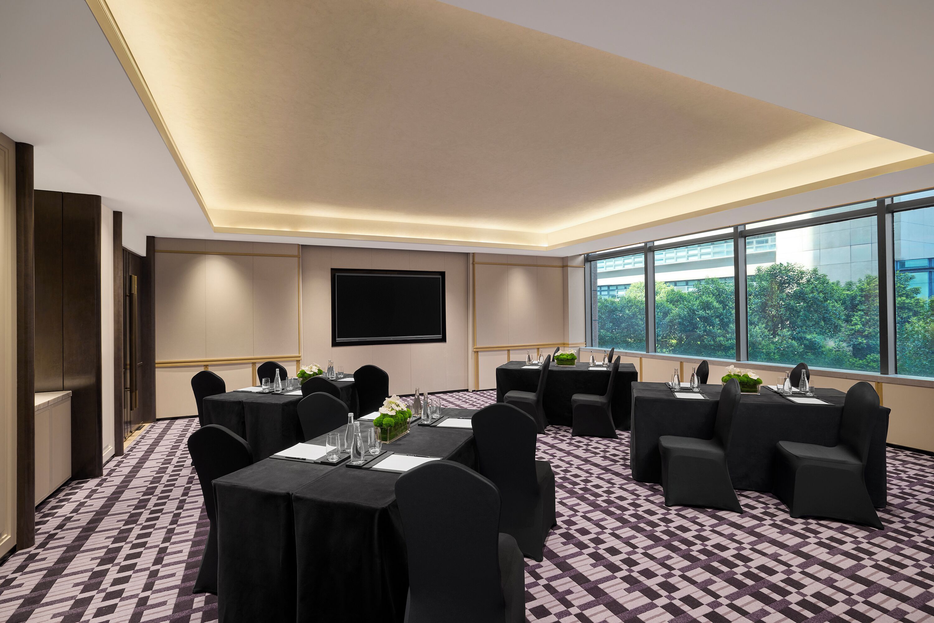 Une salle de conférence équipée de tables, de chaises, d’un écran de projection et de grandes fenêtres donnant sur un paysage verdoyant.
