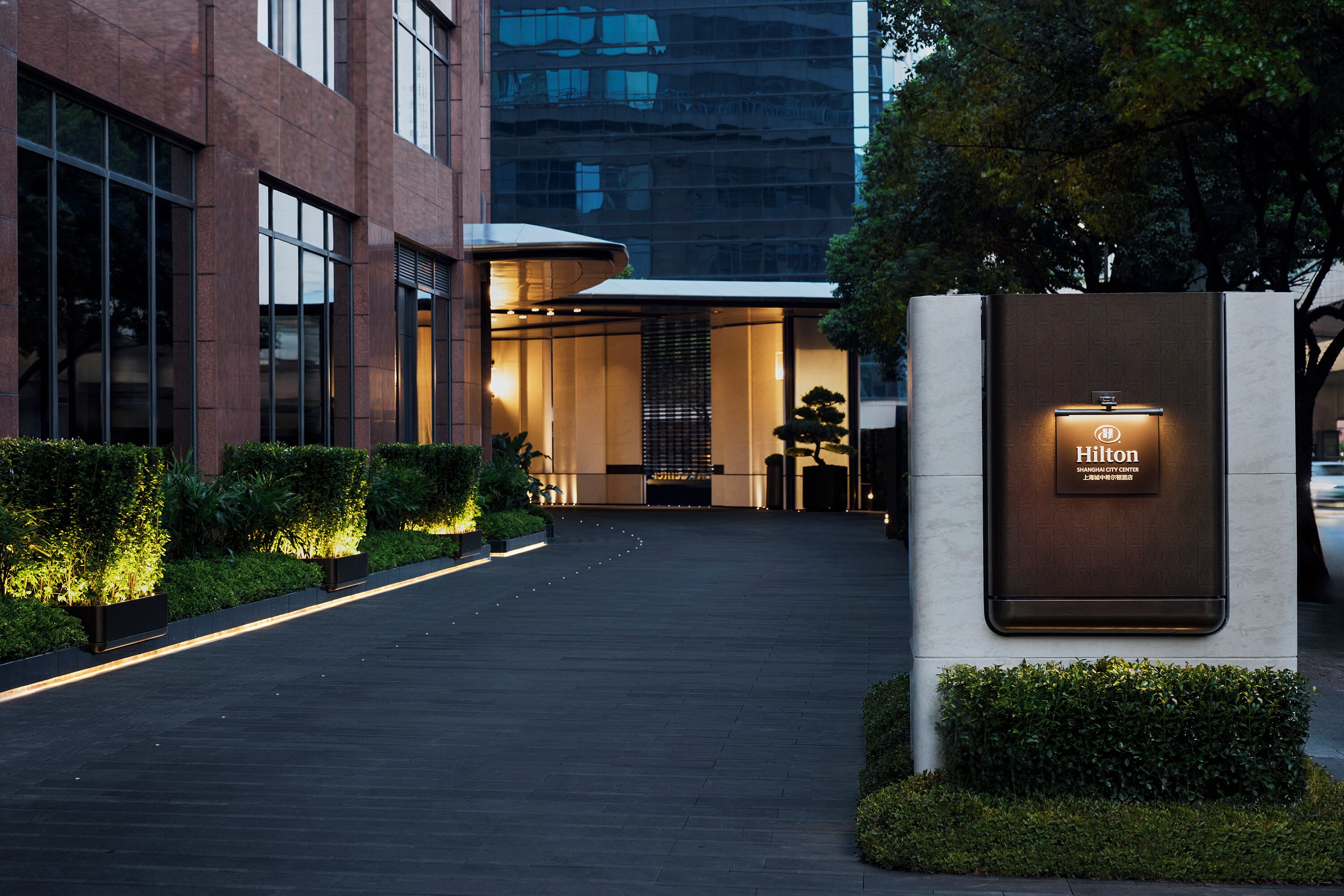 Une entrée d’hôtel moderne, avec un chemin bien éclairé, un comptoir d’accueil et une enseigne indiquant la marque Hilton.