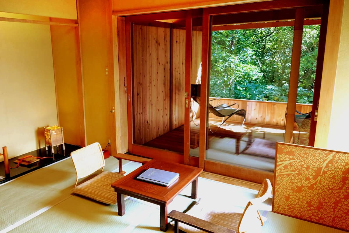 Une pièce de style japonais traditionnel, avec un sol recouvert de tatamis, une télévision, une petite table avec un livre et une porte coulissante en verre donnant sur un espace extérieur aménagé pour s’asseoir.