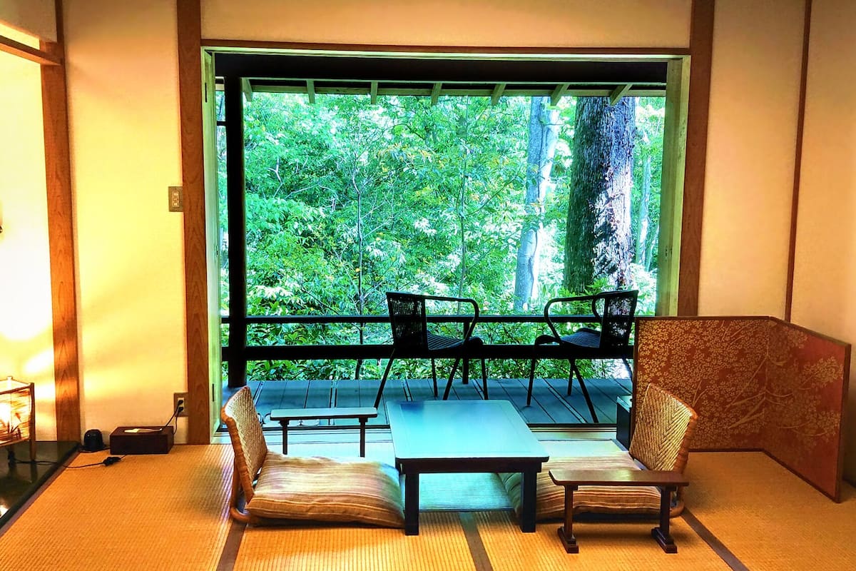 Une pièce avec vue sur une végétation luxuriante, comprenant un canapé, deux fauteuils, une petite table et une lampe décorative.