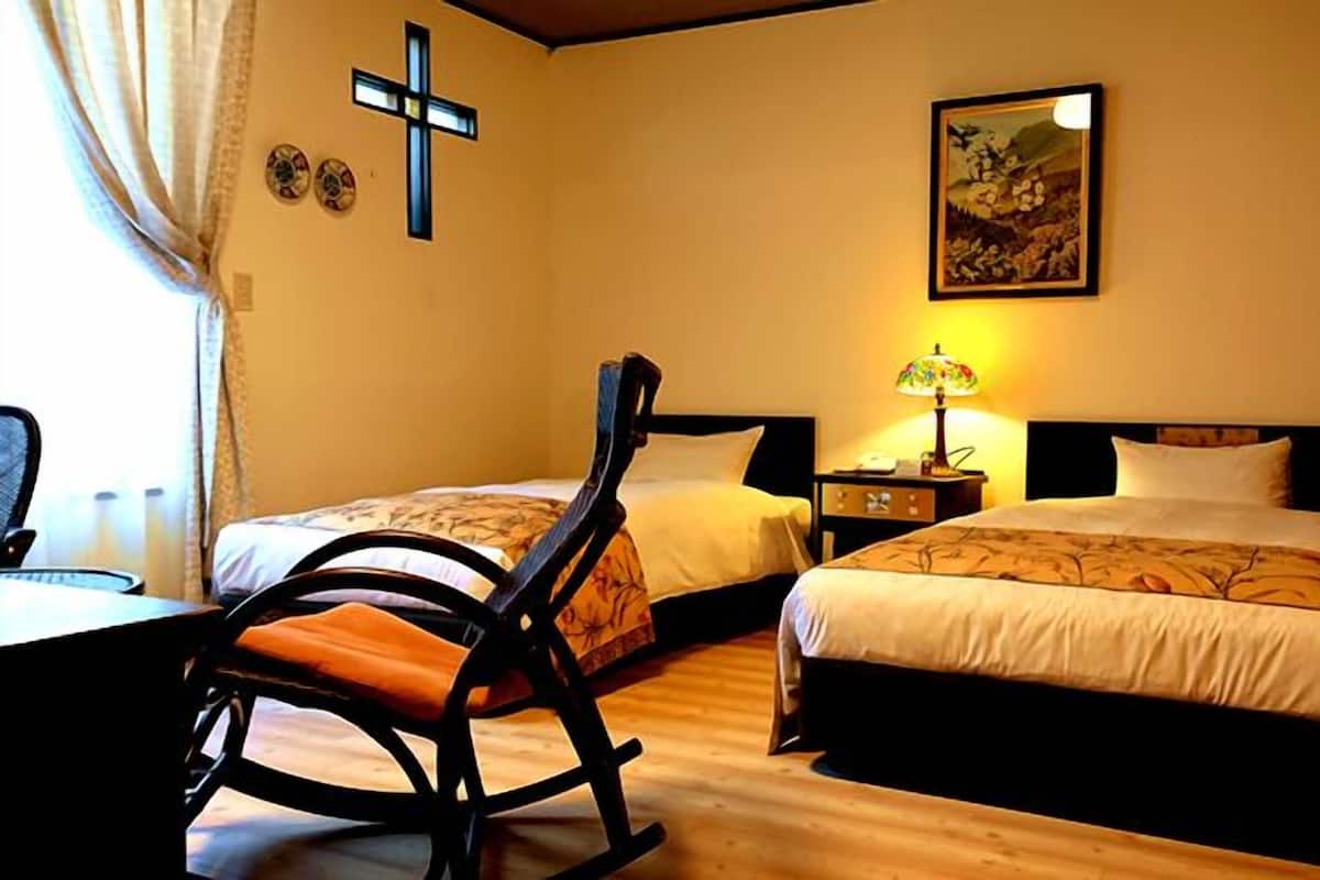 Une chambre d’hôtel avec deux lits, un fauteuil à bascule, un crucifix au mur et un tableau.