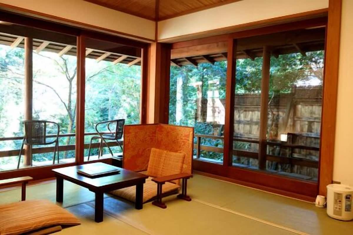 Une pièce de style japonais traditionnel, avec un sol recouvert de tatamis, une table basse, un fauteuil rembourré et de grandes fenêtres donnant sur l’extérieur.