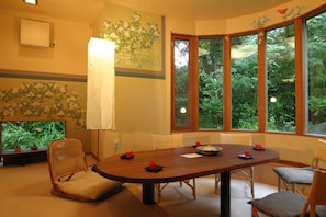 A dining area with a wooden table, chairs, and a view of greenery through large windows. - Hanafubuki (Ito)