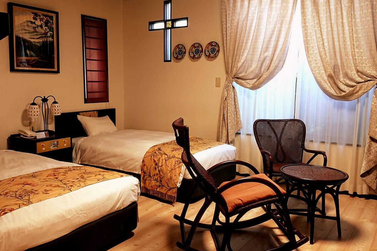 Une chambre d’hôtel avec deux lits, un fauteuil à bascule, une petite table, un crucifix au mur et un tableau.