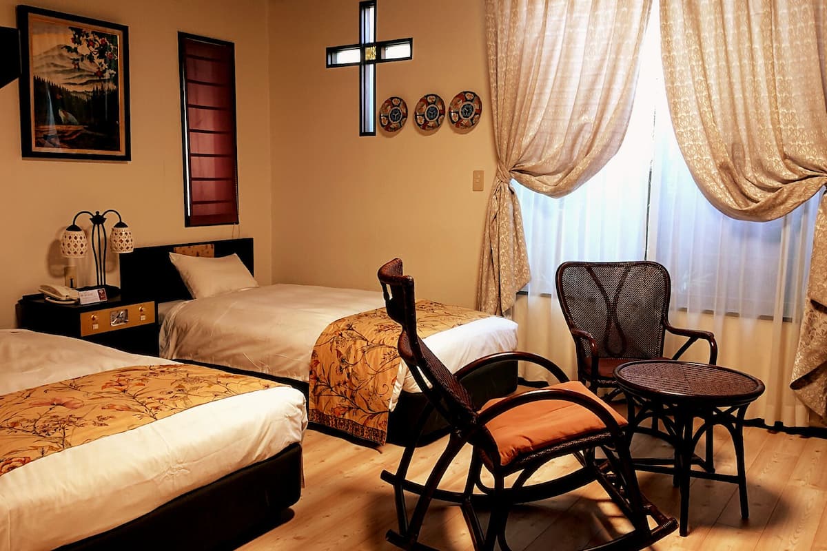 Habitación de hotel con dos camas, una mecedora, una mesita y un crucifijo en la pared, además de un cuadro.