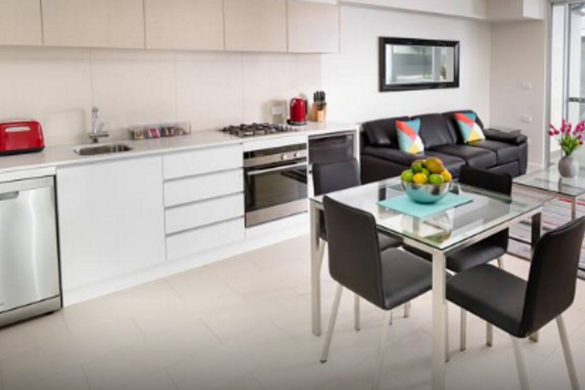 A modern kitchen with white cabinets, a dining area with a glass table and black chairs, and a sofa with colorful cushions.