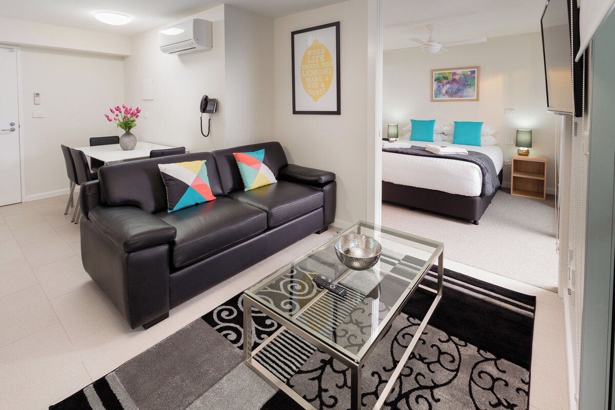 A modern hotel room with a black leather sofa, a glass coffee table, a bed with a white and gray duvet, and a small bedside table with a lamp.
