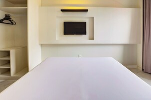 Ett minimalistiskt rum med en stor vit säng, en inbyggd TV och en hyllsektion med en klädstång.