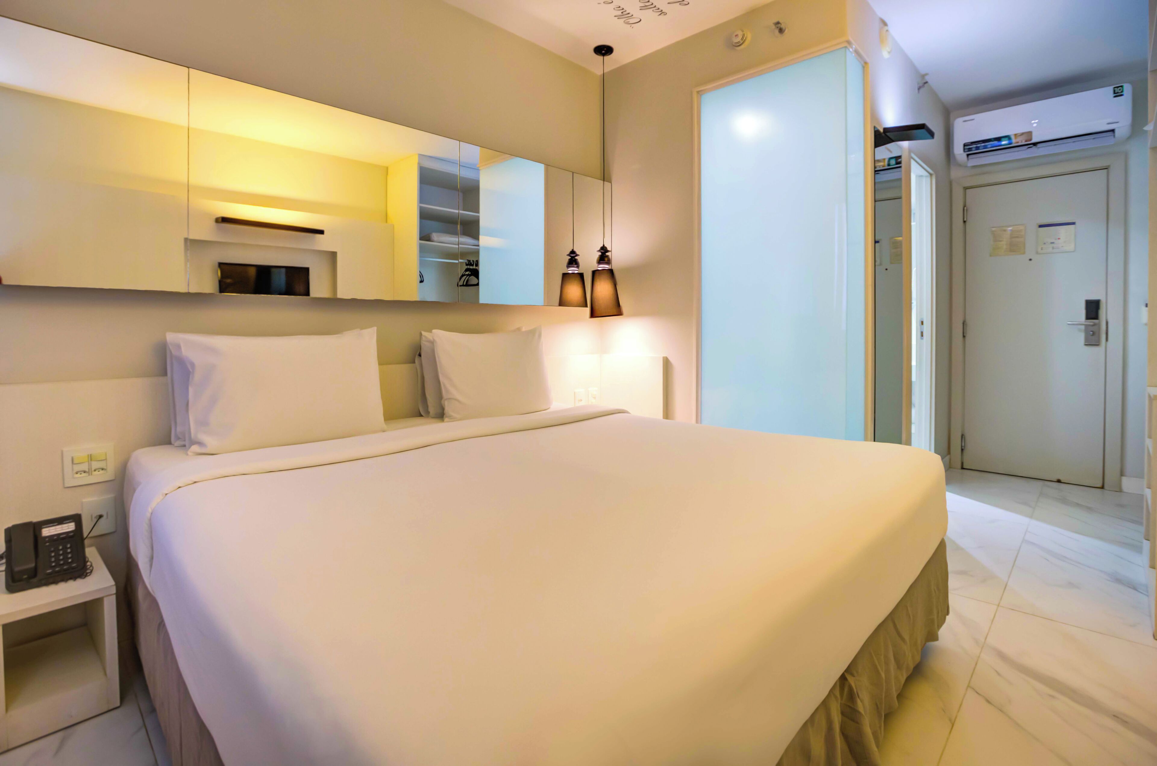 Quarto de hotel com uma cama grande, mesa de cabeceira com telefone, espelho e porta para outro espaço.