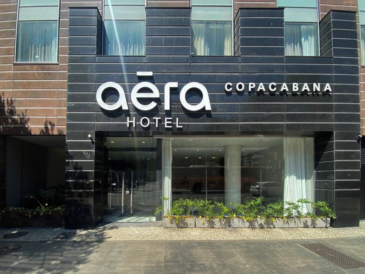 A hotel named 'Aera Copacabana' with a modern facade, large windows, and a planter with plants in front.