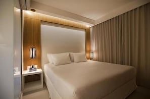 Quarto de hotel moderno com uma cama grande, mesas de cabeceira e uma cortina de um lado.