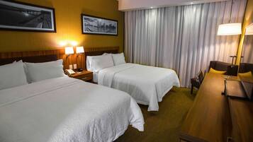 A hotel room with two beds, a wooden desk, and a large window with curtains.