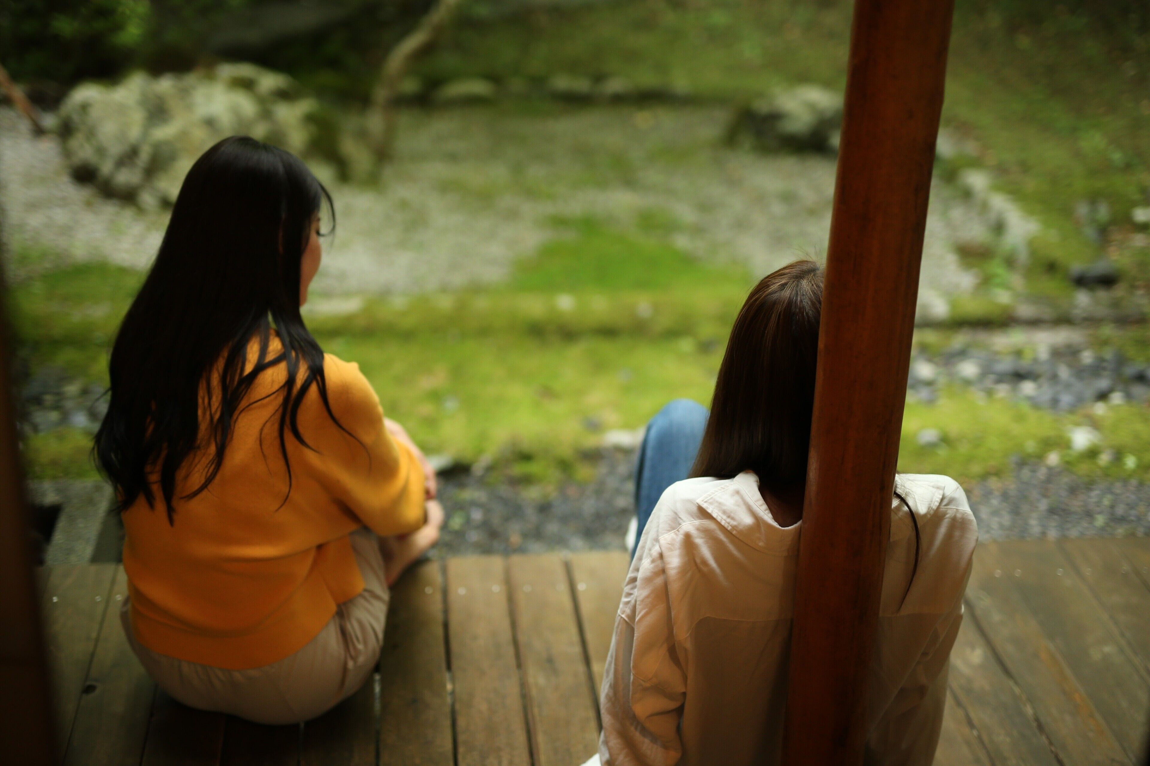 Two individuals seated on a wooden bench outdoors, overlooking a natural landscape with rocks and greenery.