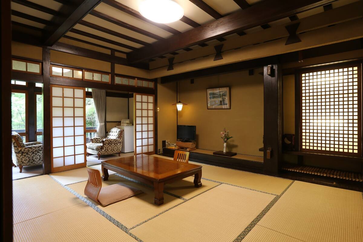 A traditional Japanese room with tatami flooring, a low wooden table, chairs, a television, and sliding doors.