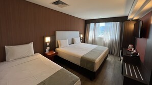 Habitación de hotel con dos camas, un escritorio con silla, televisor y ventana con cortinas.