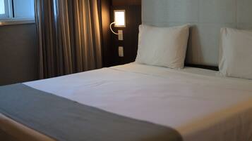 A neatly made bed with white linens, a bedside lamp, and a window with curtains.