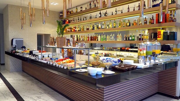 Zona de bufé con diversos alimentos y bebidas, una barra con caja registradora y estantes llenos de botellas.