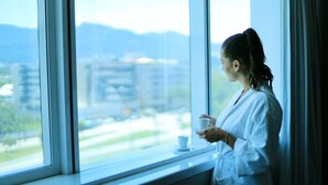 Una persona de pie junto a una ventana, sosteniendo una taza y observando un paisaje urbano con edificios y zonas verdes.