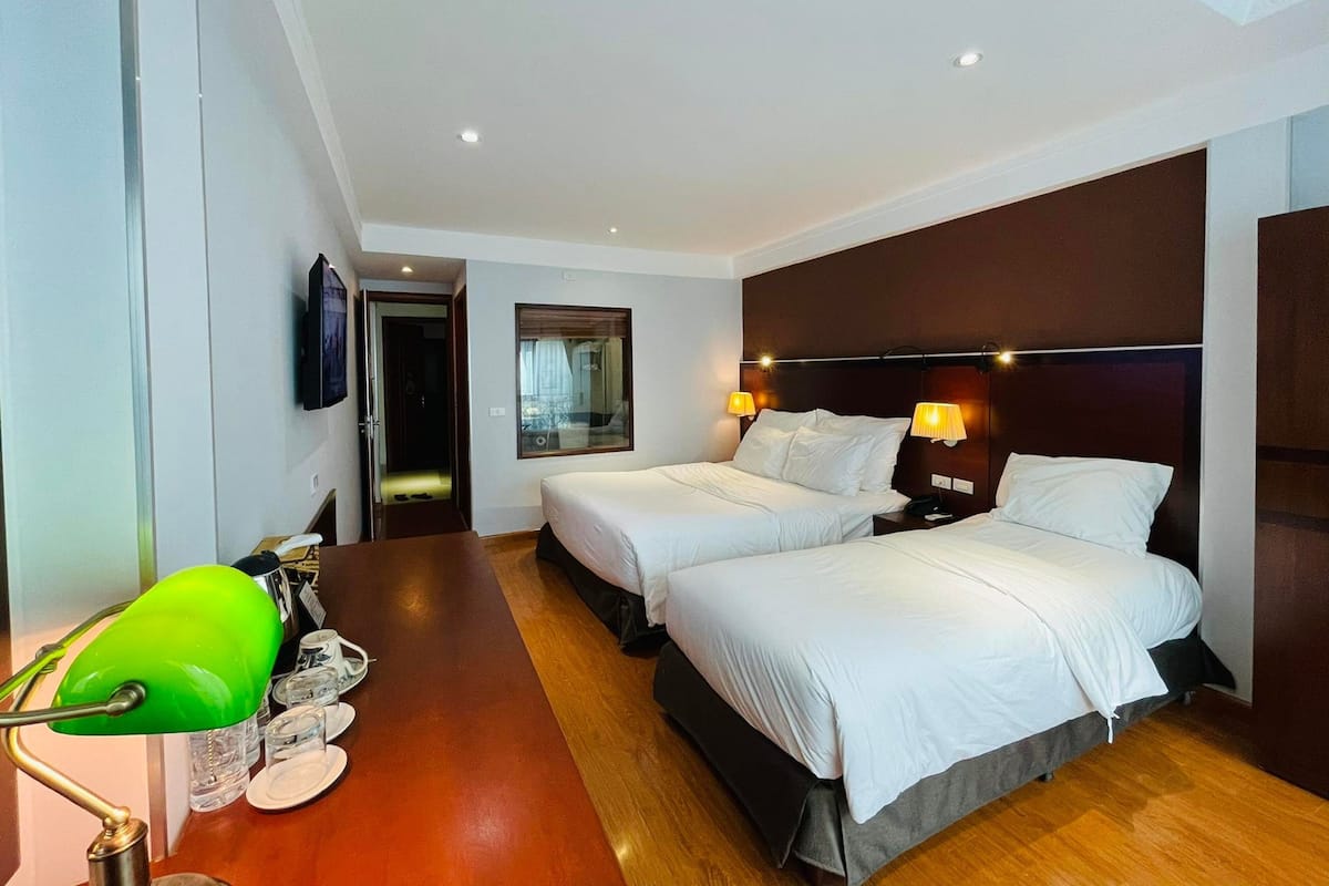 A hotel room with two beds, a desk, a green desk lamp, and a mirror.