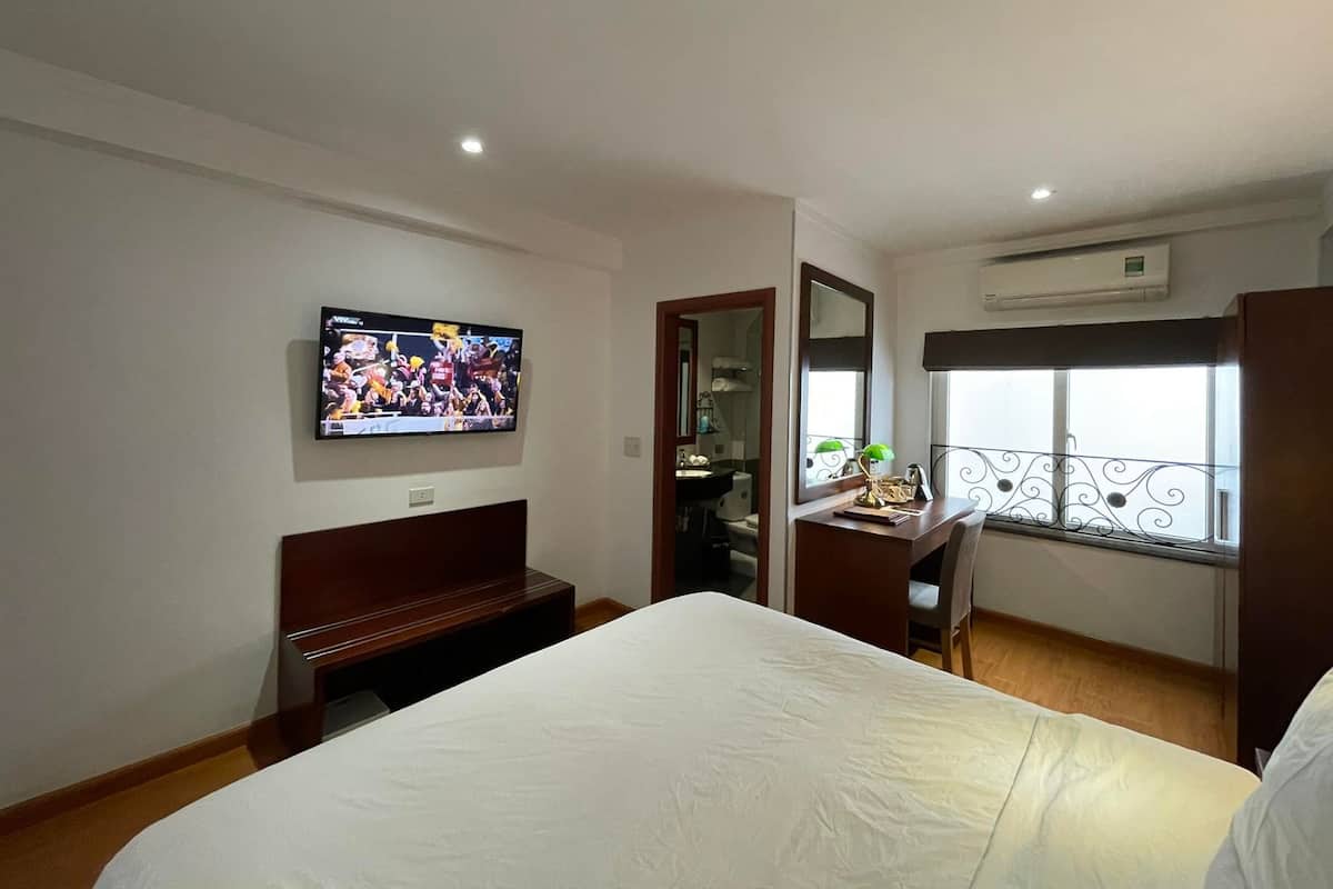 A hotel room with a bed, a desk with a chair, a television mounted on the wall, a window with a balcony, and an air conditioning unit.