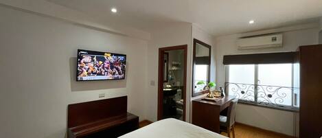 Una habitación de hotel con cama, escritorio con silla, televisor montado en la pared, ventana con balcón y aire acondicionado.