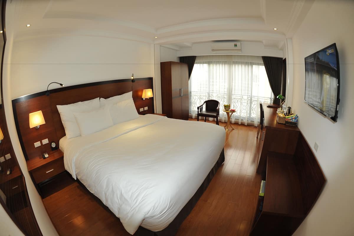 A hotel room with a large bed, wooden headboard, bedside lamps, a desk with a chair, a small table with a plant, and a large window with curtains.