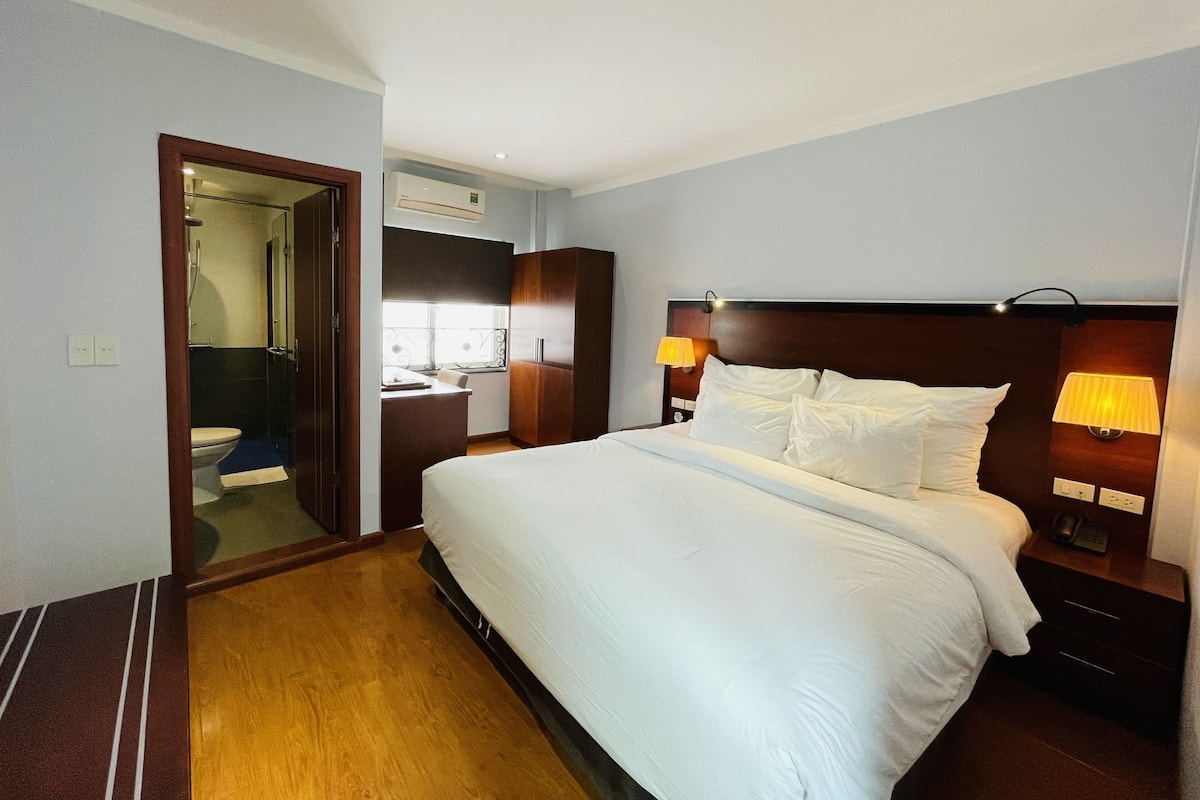 A hotel room with a large bed, bedside lamps, a wooden wardrobe, a desk with a chair, and a bathroom visible through an open door.