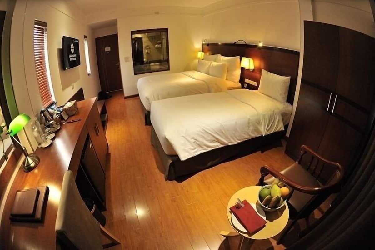 A hotel room with two beds, a desk with a lamp, a chair, and a small table with fruit.
