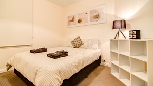 A neatly made bed with a patterned bedspread, a bedside lamp, and a wall-mounted shelf with decorative items. - Terrace on Walbourne (Launceston)
