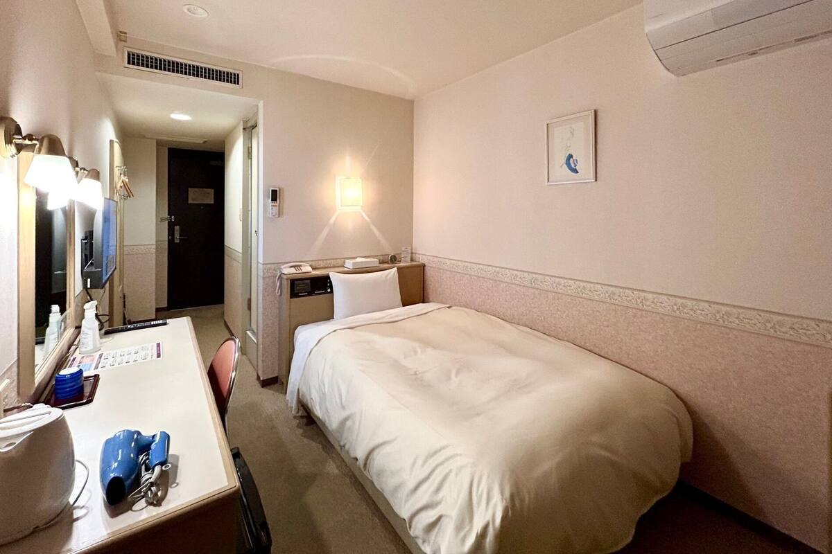 Een hotelkamer met een enkel bed, een nachtkastje met een lamp, een spiegel, een aan de muur bevestigd telefoon en een klein prentje.