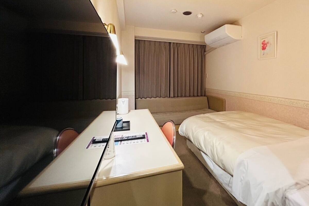 Hotelkamer met een bed, een bureau met stoel, een raam met gordijnen, een airconditioning en een klein schilderij aan de muur.