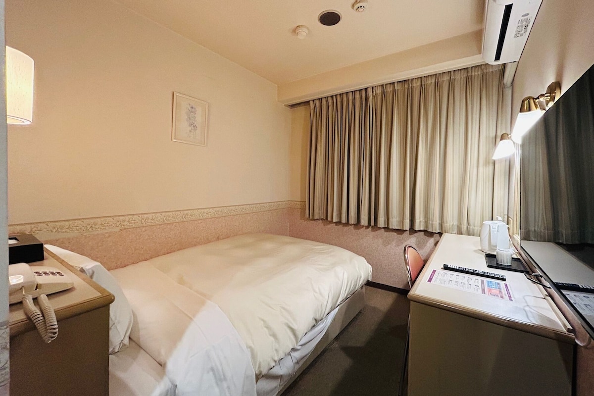 Een hotelkamer met een bed, een bureau met een computer, een telefoon en een raam met gordijnen.