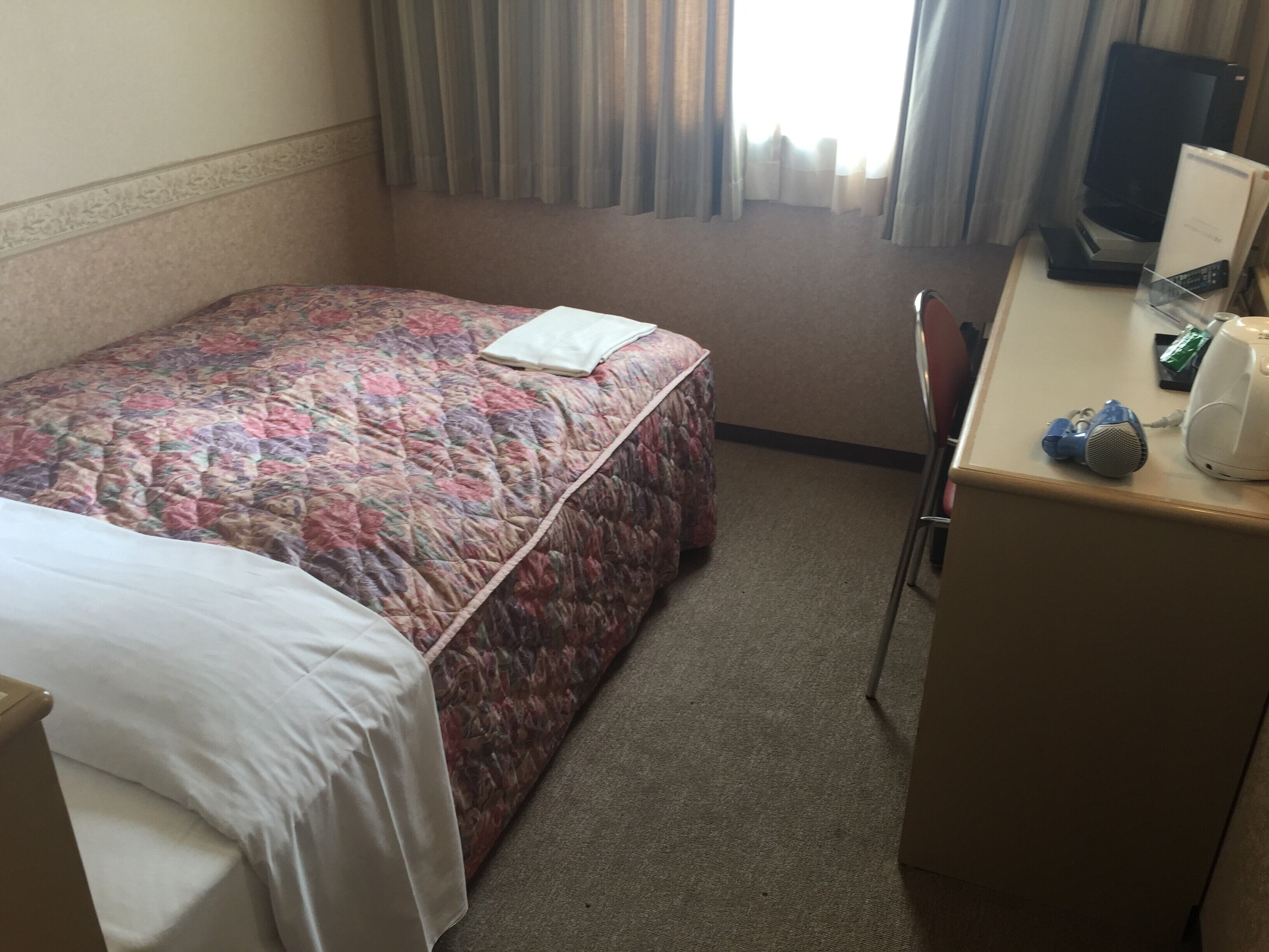 A hotel room with a bed, a desk with a computer, a chair, and a window with curtains.