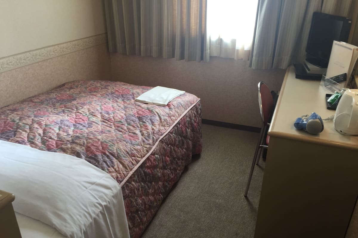 Una habitación de hotel con una cama, un escritorio con ordenador, una silla y un ventanal con cortinas.