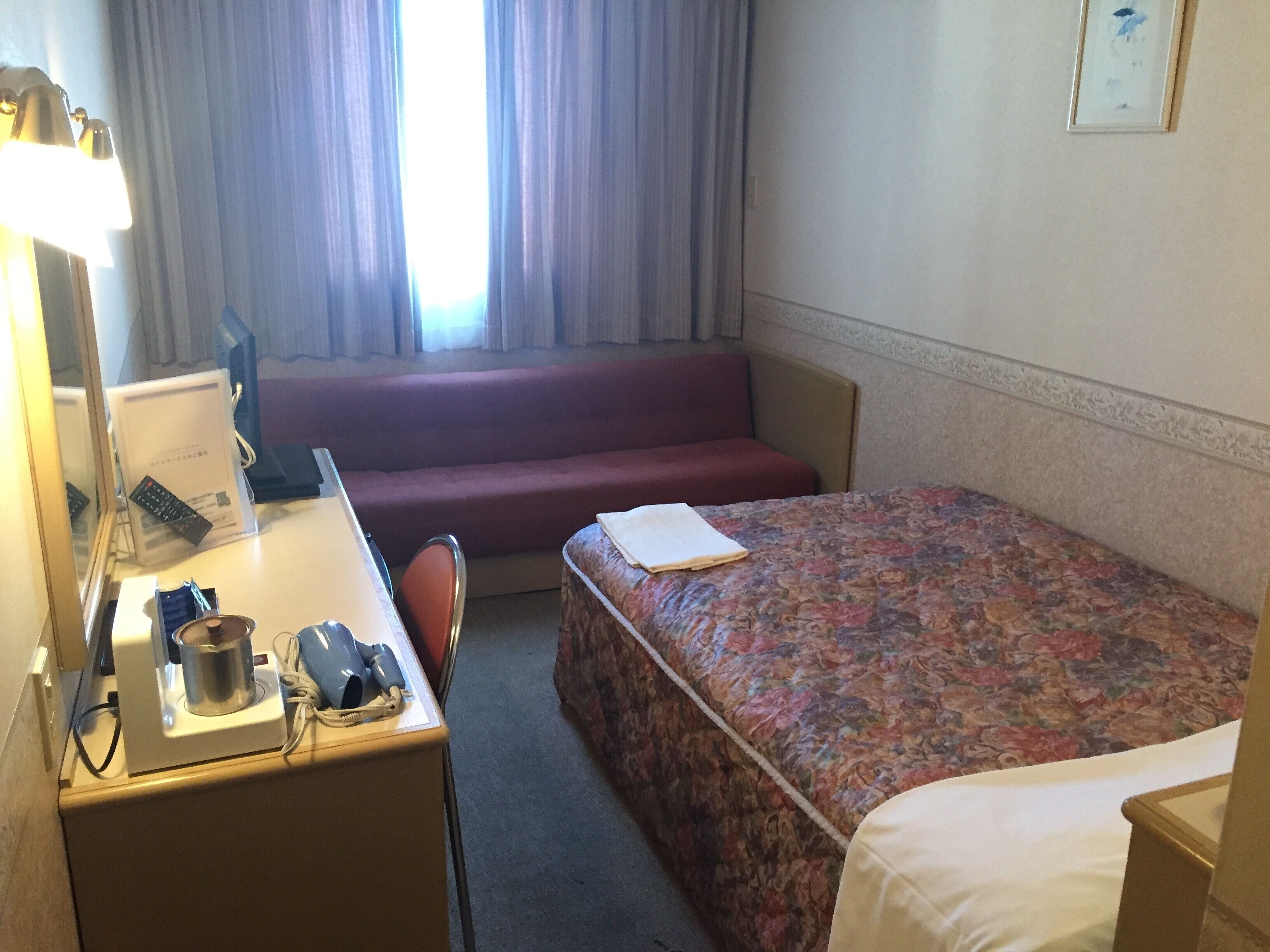 A hotel room with a bed, a desk with a lamp, a chair, a sofa, and a window with curtains.