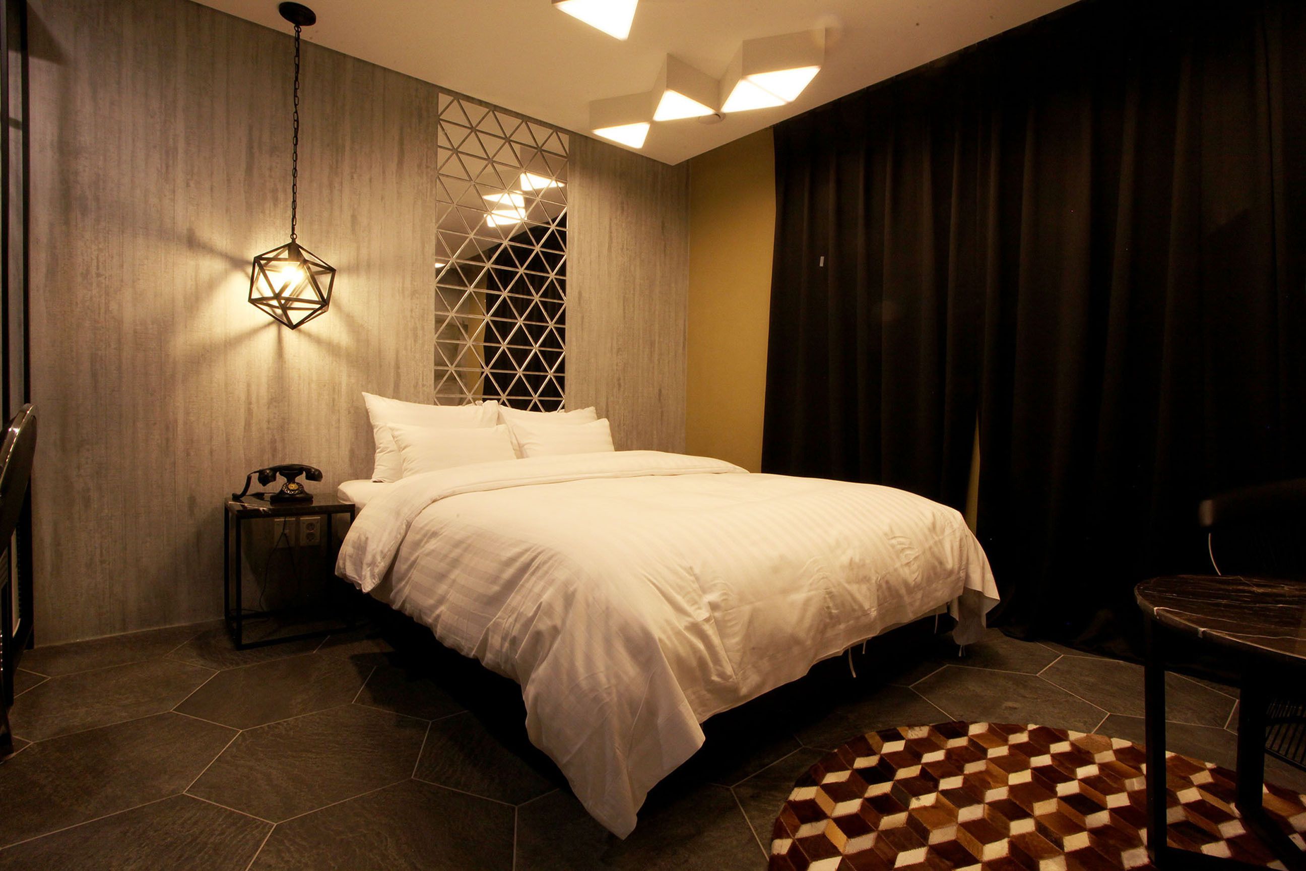 A modern bedroom with a large bed, a nightstand, a geometric-patterned rug, and a wall-mounted light fixture.
