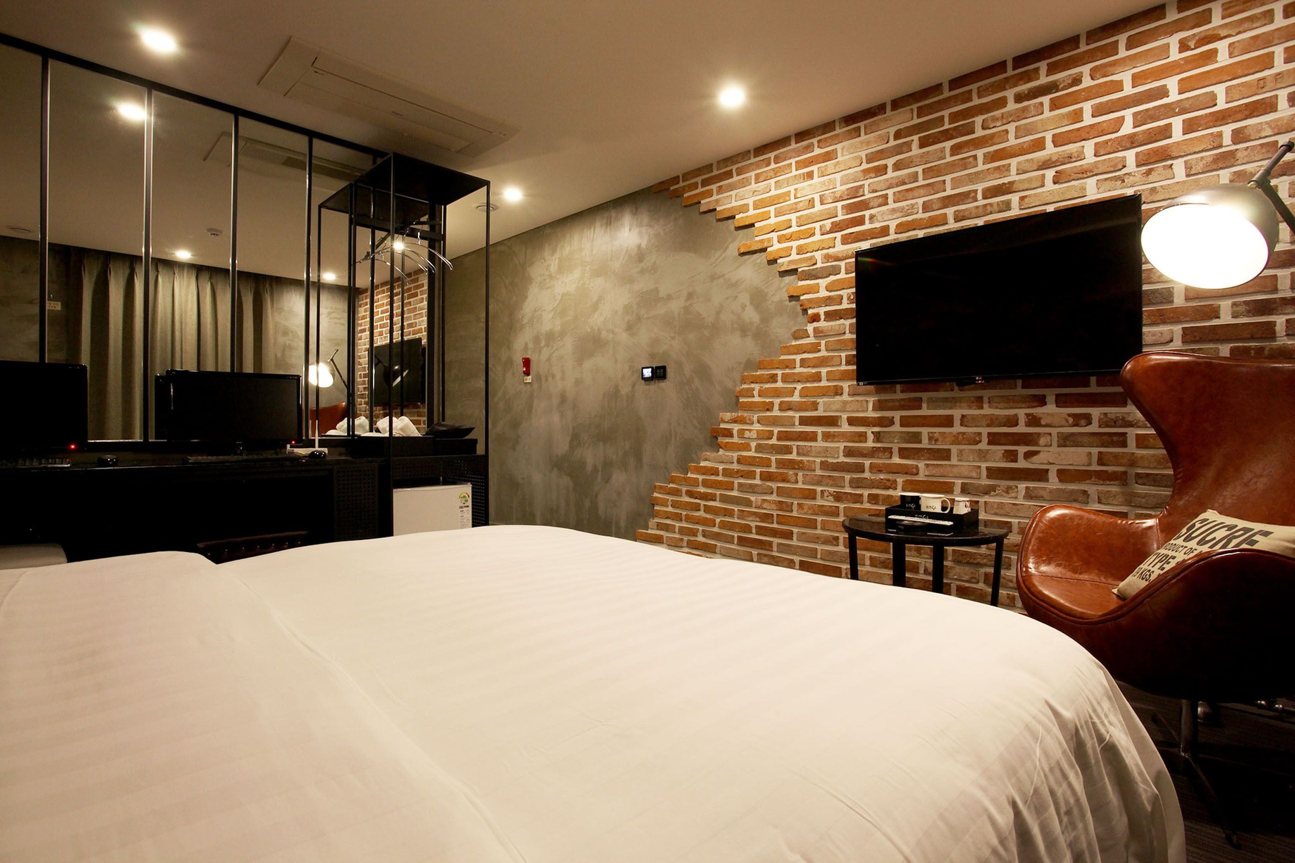 A modern hotel room with a large bed, a flat-screen TV mounted on a brick wall, a leather armchair, and a small table with a lamp.