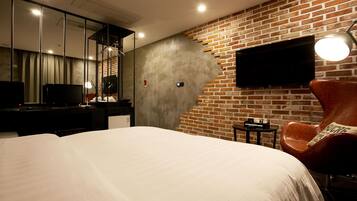 A modern hotel room with a large bed, a flat-screen TV mounted on a brick wall, a leather armchair, and a small table with a lamp.