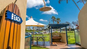 A beach-themed outdoor area with a wooden sign, a covered seating area, and a view of parked cars and palm trees. - Byron Bay Beachfront Apartments (Byron Bay)
