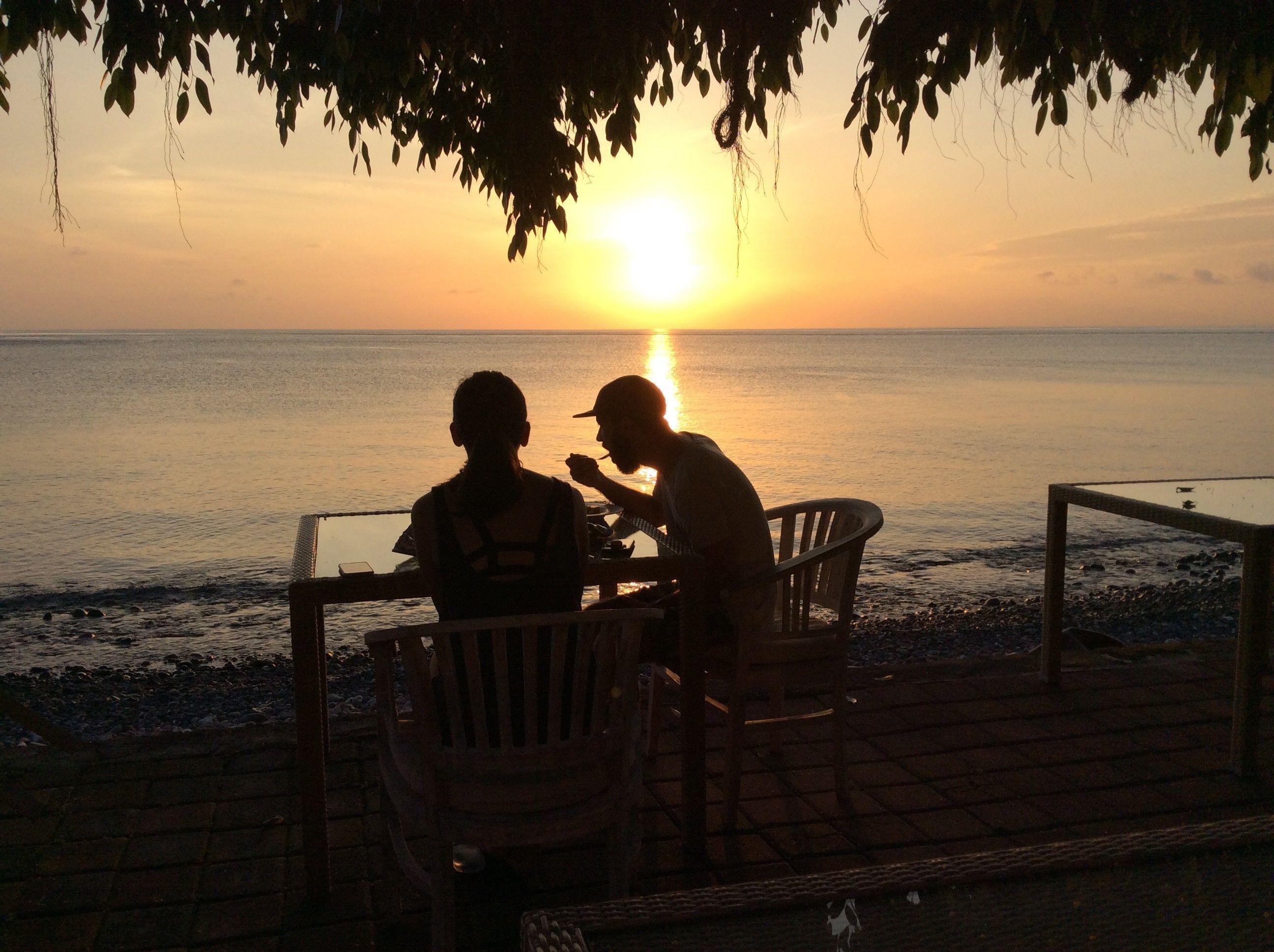 Twee personen dineren aan een tafel met uitzicht op de ondergaande zon boven de oceaan.