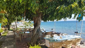 An outdoor dining area with tables and chairs, a large tree, and a view of the sea. - Nalini Resort (Karangasem)