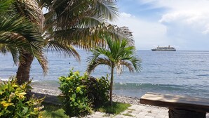 A coastal scene with a large ship in the distance, palm trees, and a bench by the water. - Nalini Resort (Karangasem)