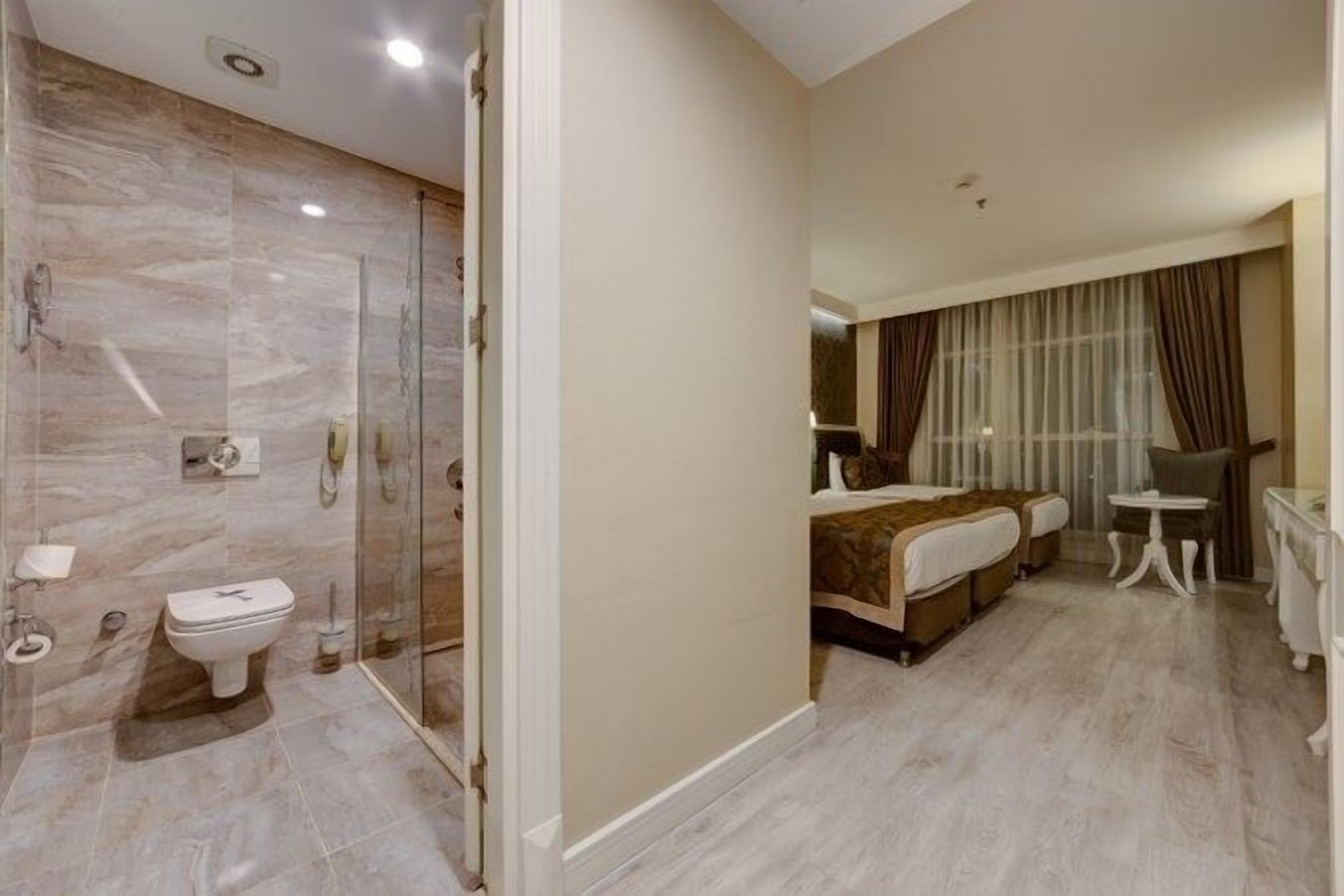 A modern hotel room with a marble bathroom, a bed with a brown headboard, a bedside table, and a large window with curtains.
