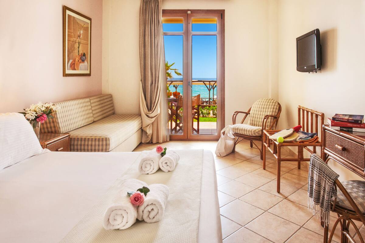 Habitación de hotel con cama, sofá, escritorio con silla, televisión y balcón con vista al mar.